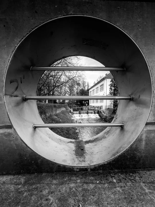 Une ouverture circulaire en béton cadre un cours d’eau et son reflet.
