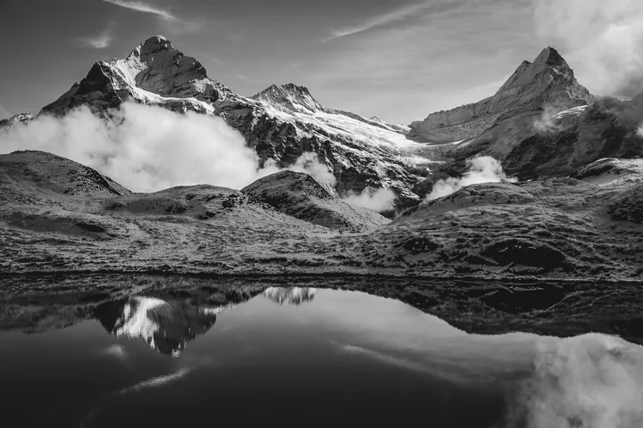 Des sommets et des nuages se reflètent dans un lac de montagne, en noir et blanc.