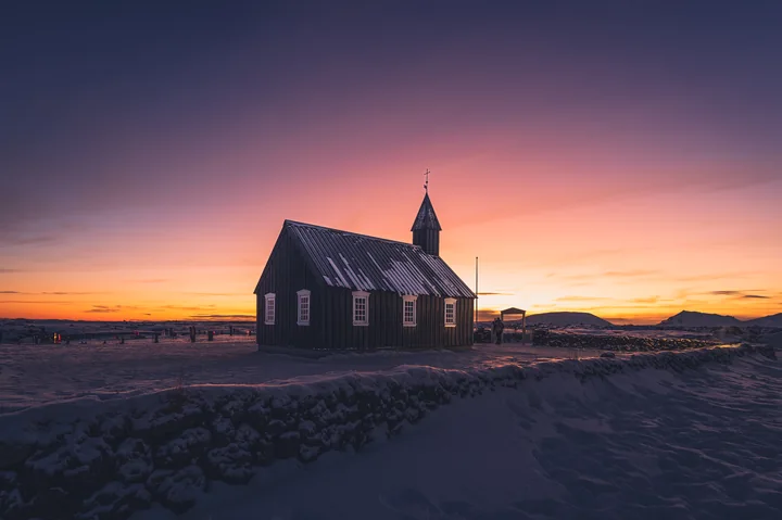 Une petite église sombre dans la neige, devant un ciel orange et violet.