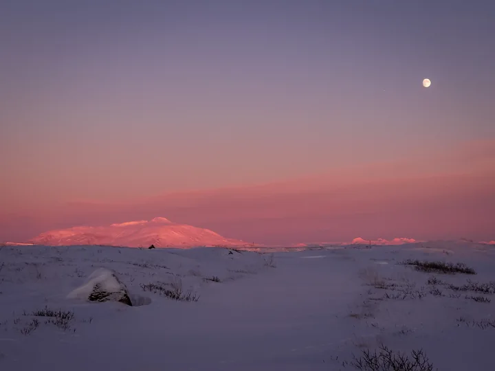 La lune se tient au-dessus de collines enneigées baignées de rose.