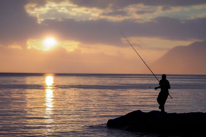 Un pêcheur en silhouette attend au bord du lac, face au soleil bas.