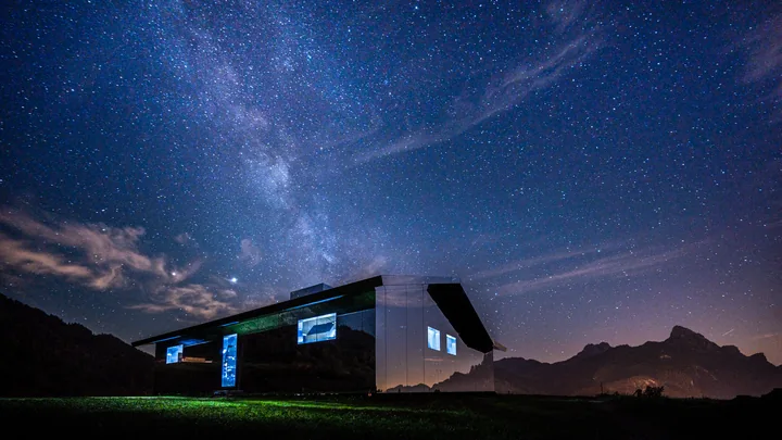 Une maison éclairée se tient sous la Voie lactée et les silhouettes des montagnes.